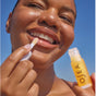 A smiling person applies OSEA Hyaluronic Lip Oil Booster with an applicator under a clear blue sky, showcasing their radiant skin and bright smile.