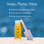 A tube of OSEA Hyaluronic Lip Oil Booster with applicator sits on sandy ground before a blurred blue backdrop. Text highlights its plumping, nourishing, glossy finish and sweet mango-mandarin scent.