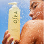 A person holds a bottle of OSEA Undaria Algae™ Body Wash in the shower, water droplets shimmering on their shoulder with a clear blue sky in the background.