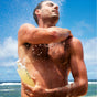 A man rinses off outdoors by the ocean, holding OSEA's Undaria Algae™ Body Wash, with blue sky and clouds in the background.