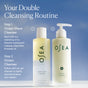 Two OSEA skincare bottles labeled “Ocean Wave Cleanser Bi-Phase Cleansing Oil and Makeup Remover” and “Ocean Cleanser” stand against a blue sky background, with text describing a two-step double cleansing routine. This regimen begins with the Ocean Wave Cleanser Bi-Phase Cleansing Oil and Makeup Remover to effortlessly dissolve waterproof makeup, followed by the Ocean Cleanser for a refreshingly thorough finish.