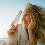 A smiling woman in a white robe holds up a bottle of OSEA Dream Night Serum outdoors, with soft sunlight and a clear sky in the background.