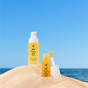 Two yellow bottles of OSEA’s Juicy Duo, Lip + Body Oil, are set on sand with the ocean and blue sky in the background—one bottle has a pump for body oil, and the other features a wand applicator for plumping lip oil.