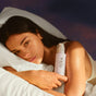 A woman with long dark hair lies on a white pillow, holding a bottle of OSEA Dream Bio-Retinol Body Serum to her chest. Softly illuminated by warm, natural light, she gazes gently at the camera.