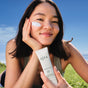 A smiling woman outdoors applies OSEA Marine Screen SPF 50 Mineral Sunscreen with non-nano zinc oxide to her cheek, holding the tube against a bright blue sky.
