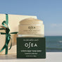 A jar of OSEA Undaria Algae™ Body Butter Bergamot & Vanilla Bean rests on its limited edition box next to a beige gift box with a green ribbon, set against a blurred ocean view under a clear sky.