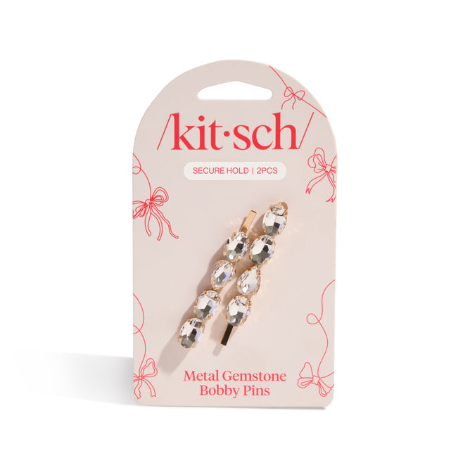 Kitsch Metal Gemstone Bobby Pins come in a pack of two gold hair pins with clear gemstones, presented on light pink packaging with red text and decorative bows for a stylish look.