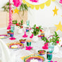 A party table set with the non-toxic Ahimsa Purposeful Plate, colorful plates, vibrant glassware, fresh fruit bowls, small flower vases, and yellow star and scalloped garlands. Pink and red accents complete the festive look.