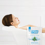 A woman relaxes in a white bathtub with her head on a towel. In the foreground, a package of San Francisco Salt Co Epsom Salts - Everyday Muscle Soak offers the ultimate muscle recovery bath.