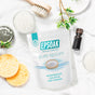 A bag of San Francisco Salt Co Pure Epsom Salts - USP Grade rests on a white surface with a yellow sponge, wooden brush, glass bottle, bowl of salt, leaves, and plate—ideal for soothing soaks and muscle relief.