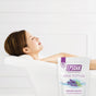 A woman relaxes in a white bathtub, eyes closed, with a bag of San Francisco Salt Co Epsom Salts - Everyday Sleep Formula featuring lavender nearby. The minimal background is entirely white.