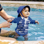 A baby in a blue sun hat and fish-patterned swim shirt stands in the pool supported by an adults hands, revealing a Green Sprouts Eco Snap Swim Diaper made from recycled polyester, as the pools edge frames the joyful scene.