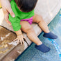 A baby sits at the edge of a pool in a Green Sprouts Eco Snap Swim Diaper made from Recycled Polyester, 18 months (22-25 lbs), sporting a green shirt, pink shorts, and navy water shoes. An adult hand supports the child as their fingers ripple through the sunny water.