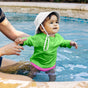 A baby in a bright green shirt and white sunhat, wearing a Green Sprouts Eco Snap Swim Diaper made from recycled polyester (18 months, 22-25 lbs), is held by an adult in a pool. The baby smiles, enjoying the calming blue water against the tiled edge.