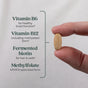 A hand holds an oval tablet of New Chapter Fermented Vitamin B Complex beside text listing USDA Organic ingredients: Vitamin B6 (supports brain health), methylated Vitamin B12, fermented biotin, and methylfolate.