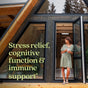 A person in a green dress gazes thoughtfully at the door of a wooden A-frame cabin. Text reads, Experience stress relief, cognitive function, and immune support with the superfood benefits of New Chapters Organic Holy Basil Booster Powder.* Large windows display trees enhancing mental wellness.