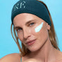 A woman wearing a dark blue RIE headband has a swipe of FRÉ Skincare's DETOX ME Instant Clearing Mask on her cheek and smiles slightly, set against a blue background.
