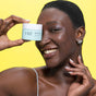 A smiling woman with short hair holds a jar of FRÉ Skincare DETOX ME Instant Clearing Mask in front of her eye against a bright yellow background. She wears a sleeveless top and multiple earrings in one ear.