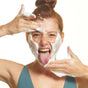 A red-haired woman with a top bun playfully applies FRÉ Skincare CLEANSE ME Foaming Micellar Water to her face, sticking out her tongue and smiling widely. She wears a teal tank top against a plain white background.