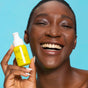A smiling person with smooth skin holds a bright yellow FRÉ Skincare C ME 10% Vitamin C Serum bottle against a blue background, highlighting its powerful skin-brightening and dark spot–reducing benefits.