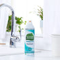 A can of Seventh Generation Disinfectant Spray sits on a white kitchen counter by a silver faucet, ready to clean nonporous surfaces, with green plants and white curtains visible in the background.