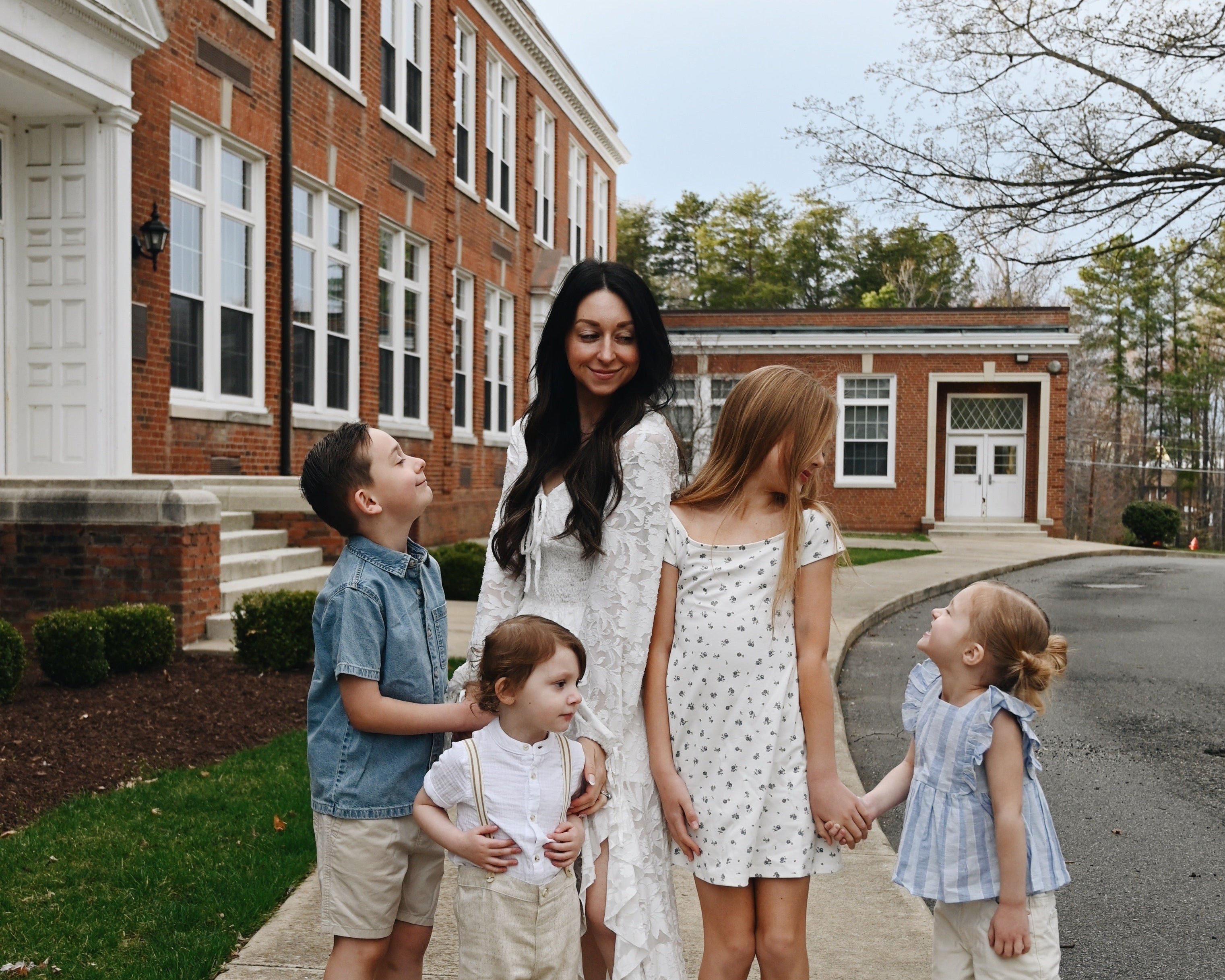 A woman in a white dress stands outside a brick building with four children. The children are smiling and interacting with each other. Trees and a sidewalk are visible in the background.