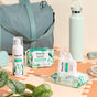 A teal gym bag, white water bottle, and a plant sit beside HoneyPot Cooling Intimate Wipes - Herbal Infusion on a peach surface, with natural light casting subtle shadows.