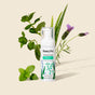 A bottle of HoneyPot Cooling Intimate Wash - Herbal Infusion rests amid fresh green herbs and a sprig of lavender on a light beige background. Its label features green botanical illustrations and showcases the cooling formula, highlighting its pH-balanced properties as an intimate cleanser.
