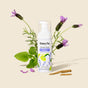 A bottle of HoneyPot Calming Intimate Wash - Herbal Infusion stands upright, surrounded by fresh green mint leaves, purple lavender flowers, and two root pieces, all set against a light beige background.