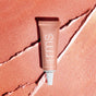 A pink tube of RMS Beauty Eyelights Cream Eyeshadow with a white nozzle cap rests on a textured, shimmery peach background featuring diagonal lines. The metallic tube is labeled “Eyelights Cream Eyeshadow.”.
