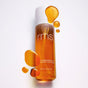 A clear bottle of RMS Beauty Kakadu Beauty Body Oil with a white cap, surrounded by floating golden oil droplets.