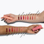 Two forearms with different skin tones each display ten swatches of RMS Beauty Legendary Lip Oil shades—Milla, Alessandra, Iris, Kaia, Lucie, Amelie, Elle, Crystal Slipper, and Adriana. Each hand holds a clean-ingredient lip oil.