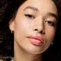 A close-up of a woman with curly hair, glossy pink lips, gold hoop earrings, and winged eyeliner. Her glowing skin stands out as she gazes confidently. RMS Beauty Legendary Lip Oil is written in the bottom left corner.