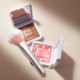 Two open makeup compacts—one with bronze powder, one with pink blush—sit on a beige surface, with the RMS Beauty Skin2Skin Powder Blush Brush lying diagonally between them for a flawless, airbrushed effect and natural look.