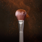 A close-up of the RMS Beauty Skin2Skin Powder Blush Brush with brown powder bursting from its bristles against a dark background, highlighting its airbrushed effect for a flawless, natural finish.
