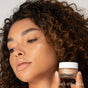 A woman with curly hair holds a small jar of RMS Beauty Master Radiance Base near her face, highlighting her radiant glow. Infused with Organic Jojoba, the formula enhances luminous skin. Rich in Radiance appears in the bottom right corner.