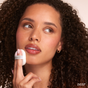 A woman with curly hair and dewy skin applies RMS Beauty’s Hydra Setting Powder to her cheek with a soft puff. She wears natural makeup, looks slightly upward, and the word DEEP appears in the bottom right corner.