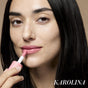A woman with long dark hair applies the hydrating RMS Beauty Legendary Lip Oil in pink, looking directly at the camera. The name KAROLINA appears in white text at the bottom right.
