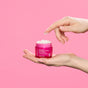 A hand holds an open jar of Andalou 1000 Roses Biome Balancing Day Cream, with a dab of the pink product on a finger.