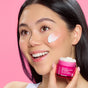 A smiling person with a dab of 1000 Roses Biome Balancing Day Cream on their cheek holds a jar of the product.