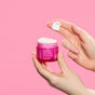 A hand holds an open jar of pink Andalou Naturals 1000 Roses Fortifying Night Cream, with a small dab of the product on a finger.