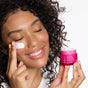 A woman with curly hair applying Andalou Naturals 1000 Roses Fortifying Night Cream to her cheek while holding an open jar of the product.