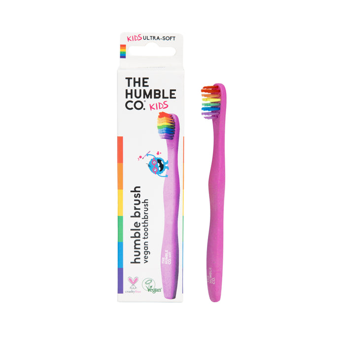 A pink Kids Rainbow Brush by The Humble Co. with ultra-soft, rainbow bristles stands by its colorful box featuring a unicorn, eco-friendly icons, and recyclable packaging.