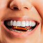 A close-up of a smiling mouth with white teeth gently holding a transparent, amber Omega-3 Fish Oil capsule from True Grace between the teeth.
