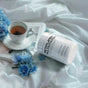 A white Codeage Methylated Multivitamin+ bottle rests on a silky surface beside tea, a book, blue flowers, and clear cubes, creating a calm, elegant scene that emphasizes essential vitamins.