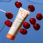 A tube of Solara Suncare Go! Mineral Defense Sport: Antioxidant Face Screen, SPF 50 rests on a reflective blue surface amid glossy red cherries. The white and orange tube has a yellow design and displays the SPF 50 label.