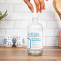 A hand places a spray nozzle into a clear bottle of Meliora All-Purpose Home Cleaner that contains All-Purpose Home Cleaner Refill Tablets, on a kitchen counter with white tile backsplash and blue-patterned containers.