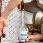 A person fills a Meliora All-Purpose Home Cleaner bottle with water from a kitchen faucet while another hand holds the bottle. In the background, a dish rack and dishes highlight how convenient it is to use Meliora All-Purpose Home Cleaner Refill Tablets for your cleaning spray routine.
