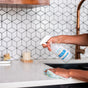 A person sprays Meliora All-Purpose Home Cleaner Refill Tablets solution onto a white kitchen countertop, holding a cloth in the other hand. The backsplash features a geometric white tile pattern, and there is a copper faucet in the background.