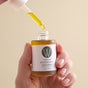 A hand holds Wildcraft’s Regenerate Face Serum, a moisturizing facial oil, using a dropper to dispense its golden-yellow liquid. The background is plain beige.